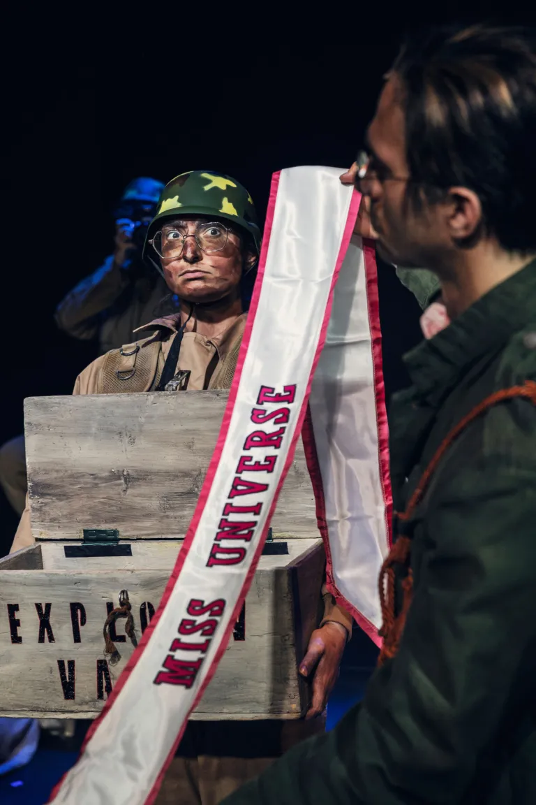 En person i militärhjälm tittar upp bakom en trälåda med ett band där det står “Miss Universe”.