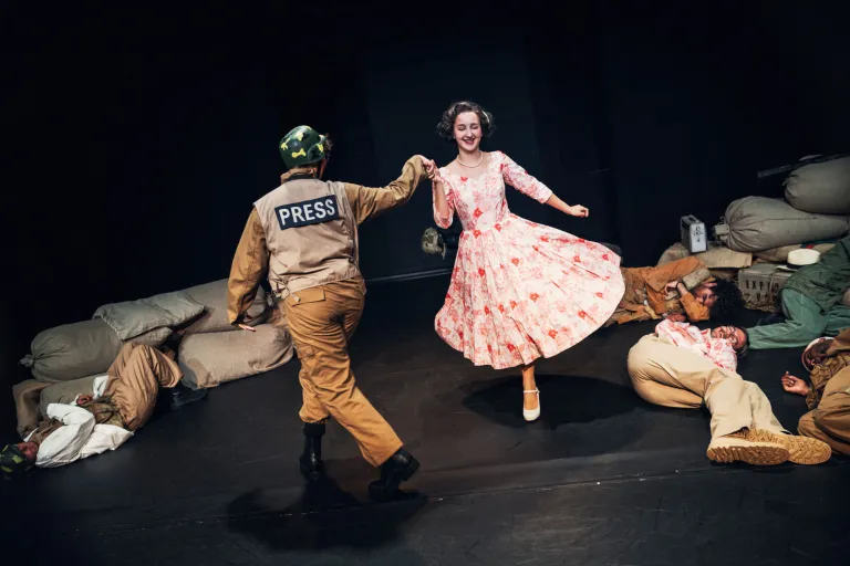 En person i blommig klänning dansar med en soldat, medan flera personer ligger utslagna på scengolvet.