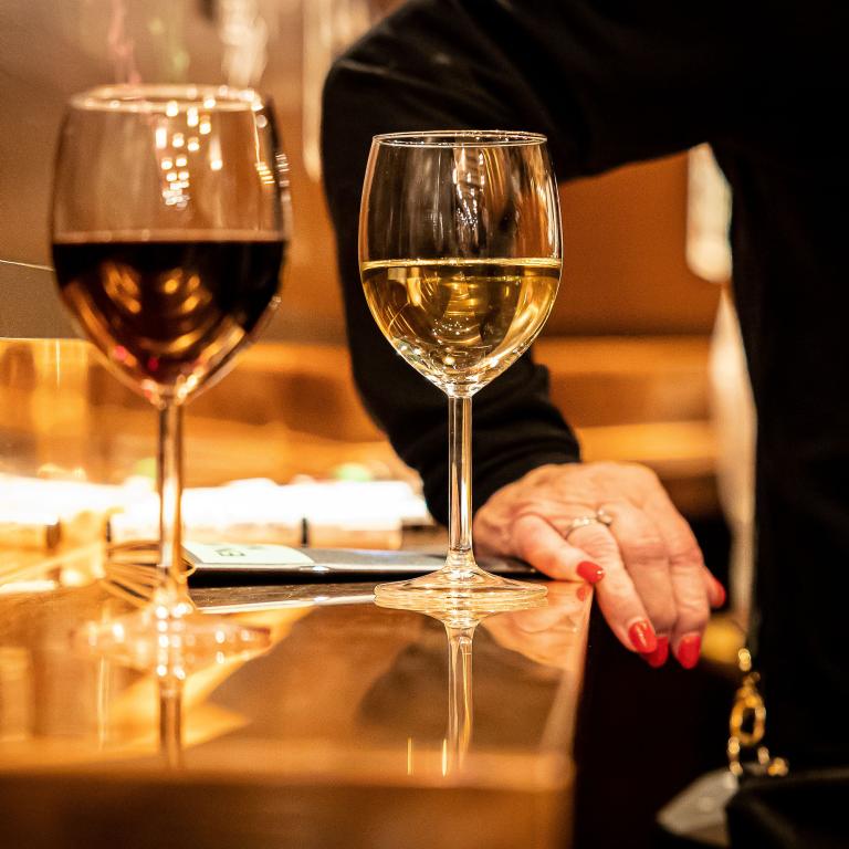 Två glas vin på en bardisk, ett rött och ett vitt. En hand med röda naglar i bakgrunden. 