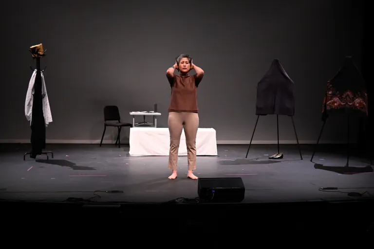 En barfota skådespelare står mitt på scenen med händerna mot huvudet, omgiven av klädställningar och rekvisita.