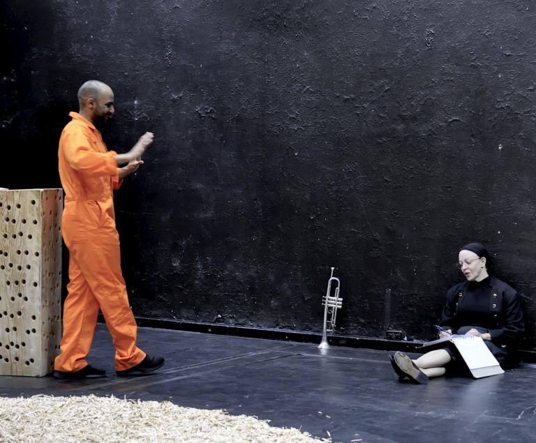 En skådespelare i svart sitter ner till höger. en skådespelare i orange dräkt står till vänster.