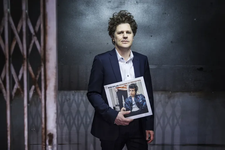 Erik står vid en industrihiss i blå snygg kostym och håller sitt favoritalbum Highway 61 Revisited