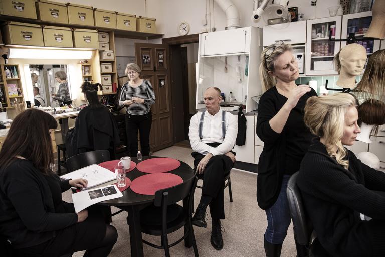 Inne i kostymavdelningens verkstad blir två skådespelare sminkade samt att frissyrer skapas medan en skådespelare sitter på en stol mitt irummet och väntar på sin tur.