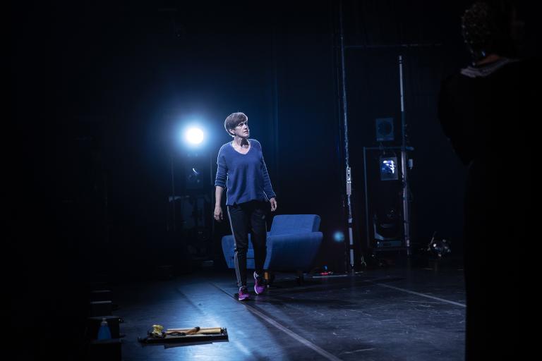 En skådespelare i privata kläder står på scenen, framför en blå fåtölj och med ett lysande blått strålkastarljus bakom sin axel. Hon tittar på något vi inte ser. Rekvisita ligger på scenen och bilden går i svarta och blå toner.