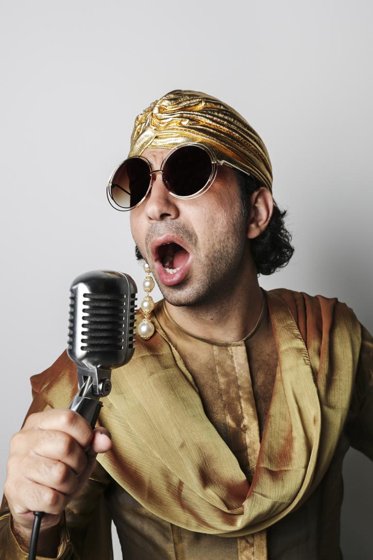 Man klädd i gulfärgade klädder och mössa håller en 50-tals mikrofon i handen och sjunger och har svarta runda solglasögon på sig samt en lång örhänge med många pärlor. 
