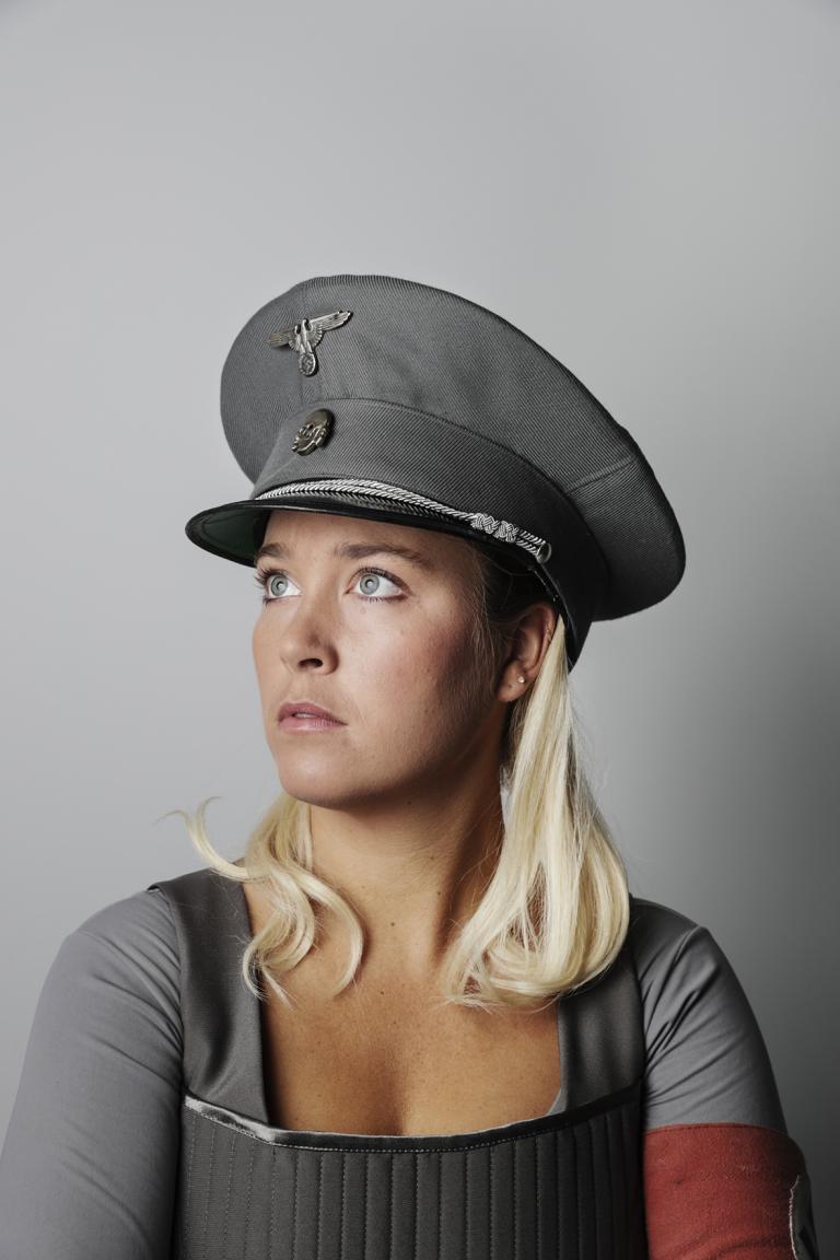 kvinna med axellångt blont hår har en mörkgrå uniformsmössa och frånvarande blick. 