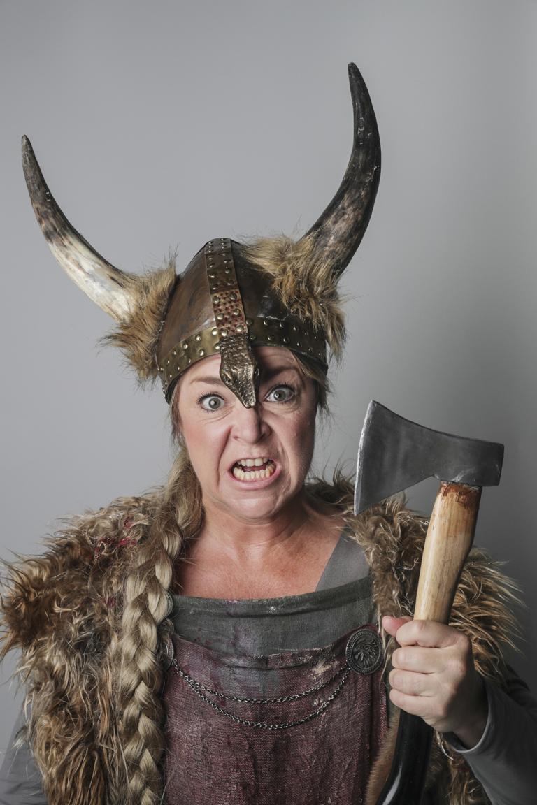 Kvinna iklädd i vikinga hjälm med horn. Hon har en arg blick och en lång fläta över axeln. Hon håller en blodig yxa i handen. 