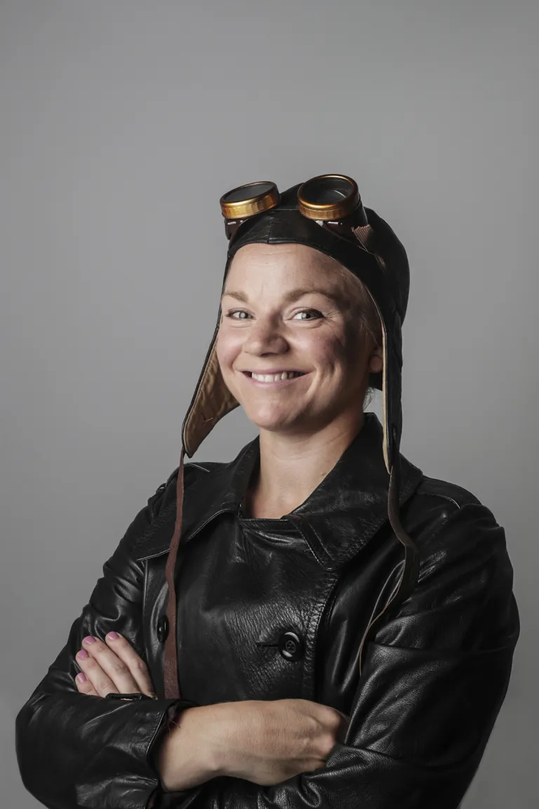 Kvinna klädd i svart skinnjacka med skinnhätta och svarta flyg solglasögon på pannan. Hon ser glad ut och självsäker.  