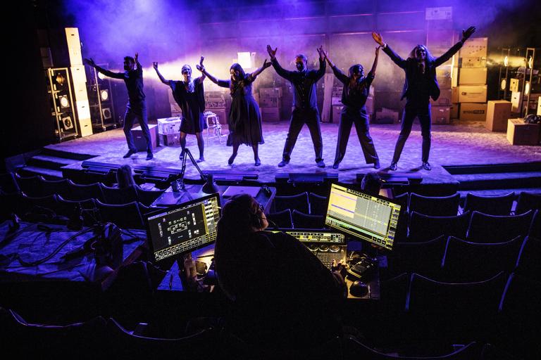 Sex personer håller sig hand i hand och dansar på teaterscenen. De är iklädda i mörka kläder och det finns en stark blå belysning över hela scenen. 