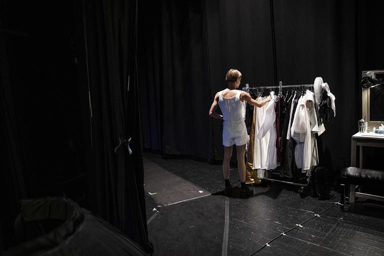 En skådespelare står och väljer kläder från ett klädställ bakom scen.