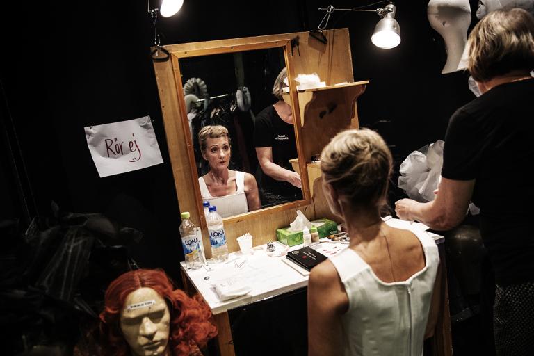 En skådespelare sitter framför en spegel och får hjälp att byta peruk