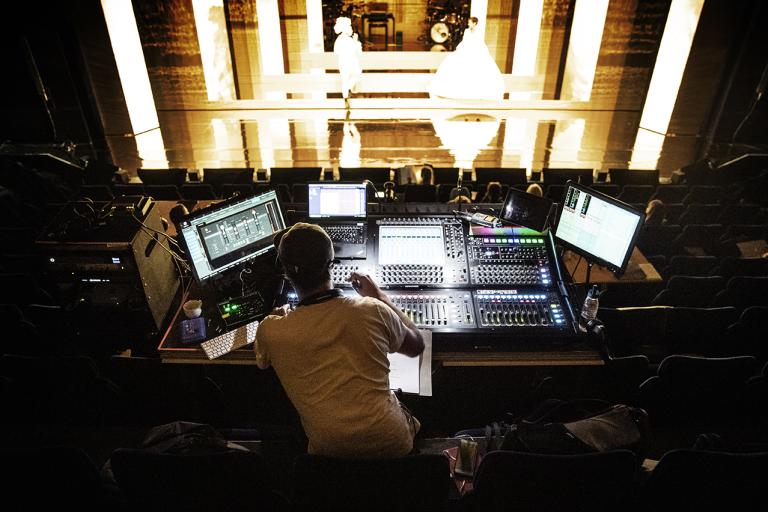 Ett stort mixerbord med lysande lampor manövreras av en ljudtekniker mitt bland publikplatserna framför scenen.