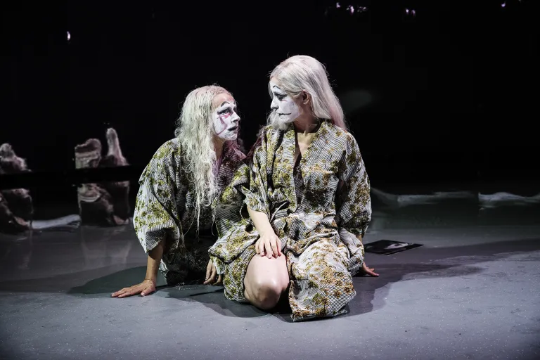 Två kvinnor sitter på golvet. Ansiktena är målade i vit mask.
