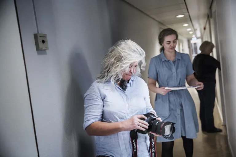 Fotografen Tine står i en korridor med sin kamera och i bakgrunden syns Stine Martinsen.