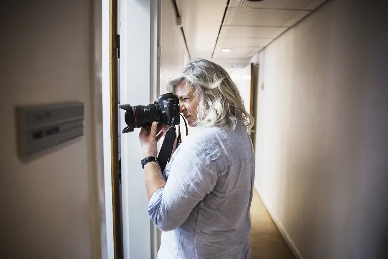 Tine Harden står i en korridor och fotograferar det som händer innanför en av dörrarna.