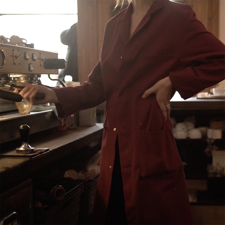 Kvinna i röd rock står vid en kaffemanskin och gör kaffe på ett Café.