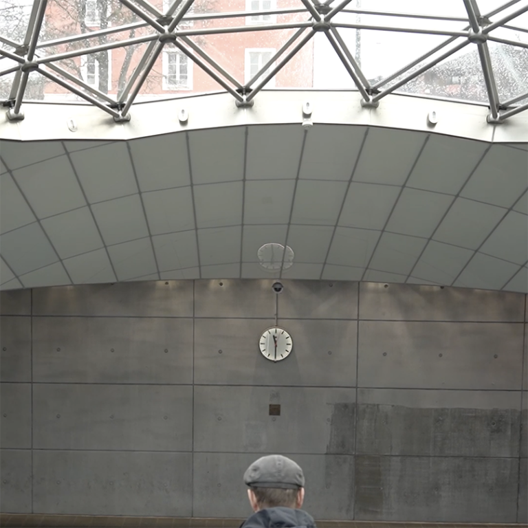 Nedgången till Triangelns tågstation. På bilden syns bakhuvudet på en man i keps som åker ner för rulltrappan och ovanför hans huvud syns en klocka.
