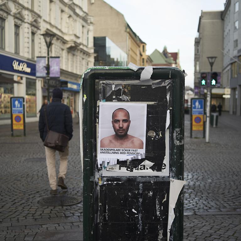 Gågatan i Malmö. På ett elskåp sitter en affisch med en skådespelare. På affischen står texten "Skådespelare söker fast jobb med pension"