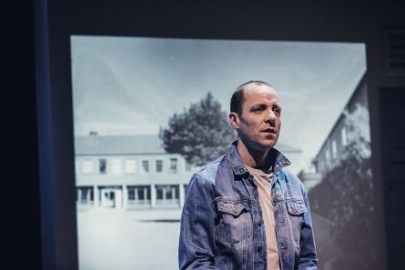Man i jeansjacka står framför en projiceringen av en svart vit bild på en skolgård