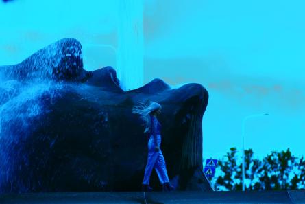 Kvinna går på kanten av en fontän och i bakgrunden syns skulpturen av ett huvud som sprutar vatten