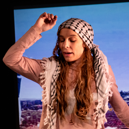 Kvinna med shemagh (palestinasjal) på huvudet och rosa tröja höjer en hand i luften i en rörelse likt att hon dansar
