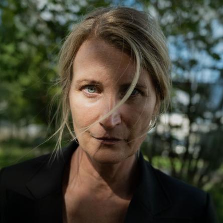 Monica Wilderoth ser in i kameran, en hårslinga framför en ögat, gröna löv i bakgrunden