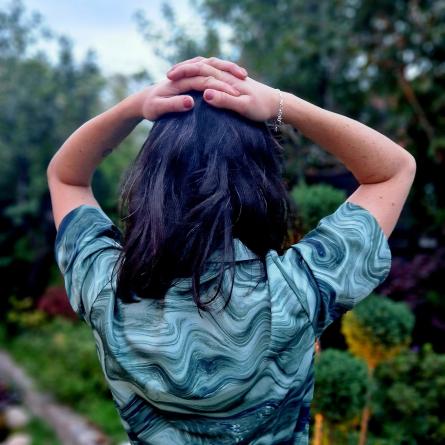 En person står i en trädgård med ryggen mot kameran, med händerna på huvudet