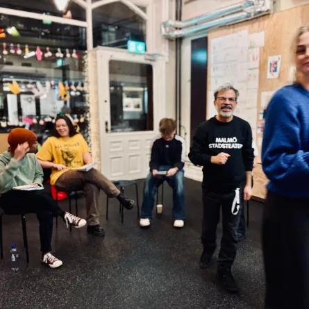 En workshopmiljö med människor i samtal och aktivitet. En man i en Malmö Stadsteater-tröja står i mitten och ler mot kameran, medan andra deltagare sitter på stolar i bakgrunden, pratandes eller skrivandes. En kvinna rör sig framåt med en anteckningsbok i handen.