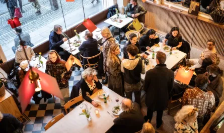 Folk sitter och står tätt kring dukade bord i en restaurangmiljö med svartvitrutigt golv.