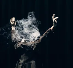 Person i svart kavaj med ryggen mot kameran står med händerna i luften och ur kavajen kommer det rök.