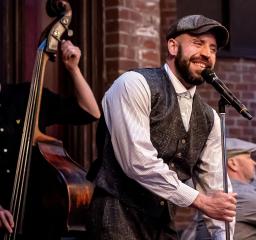 Person med skjorta, väst och keps står vid mikrofon och kontrabas i bakgrunden