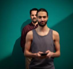 En kille med skägg och linne i förgrunden med en mobil i handen, ur fokus, i bakgrinden en äldre man som tittar på killen. 