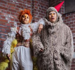 Två personer står mot en tegelvägg - en i grå pälskostym och näbbliknande strut på huvudet, en med vitt krås, vita fjädrar och små gosedjursankungar längs med benen.