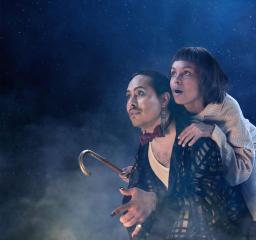 Två personer mot en stjärnhimmel, tittar bort i fjärran. En med trollkarlsliknande kostym med fluga käpp och svartguldig rock, en i ljus pyjamas.