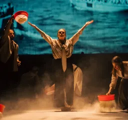 En skådespelare står mitt på scenen med armarna utsträckta medan andra håller röda hinkar i rökigt scenljus.