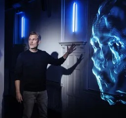 En person tittar på något utanför bild och pekar mot en digital projicering av ett ansikte