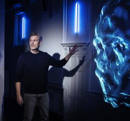 En person tittar på något utanför bild och pekar mot en digital projicering av ett ansikte