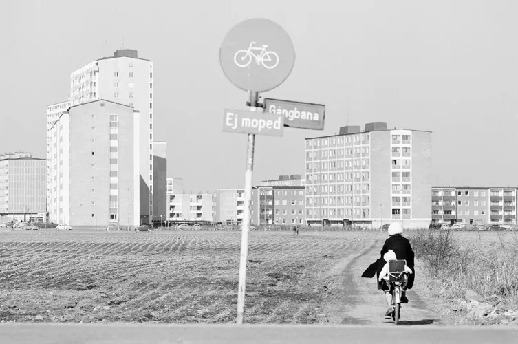 svartvit bild över ett fält och en person som cyklar mot en grupp nybyggda flervåningshus. I förgrunden syns en vägskylt med cykel och texterna "Gångbana" och "Ej moped"