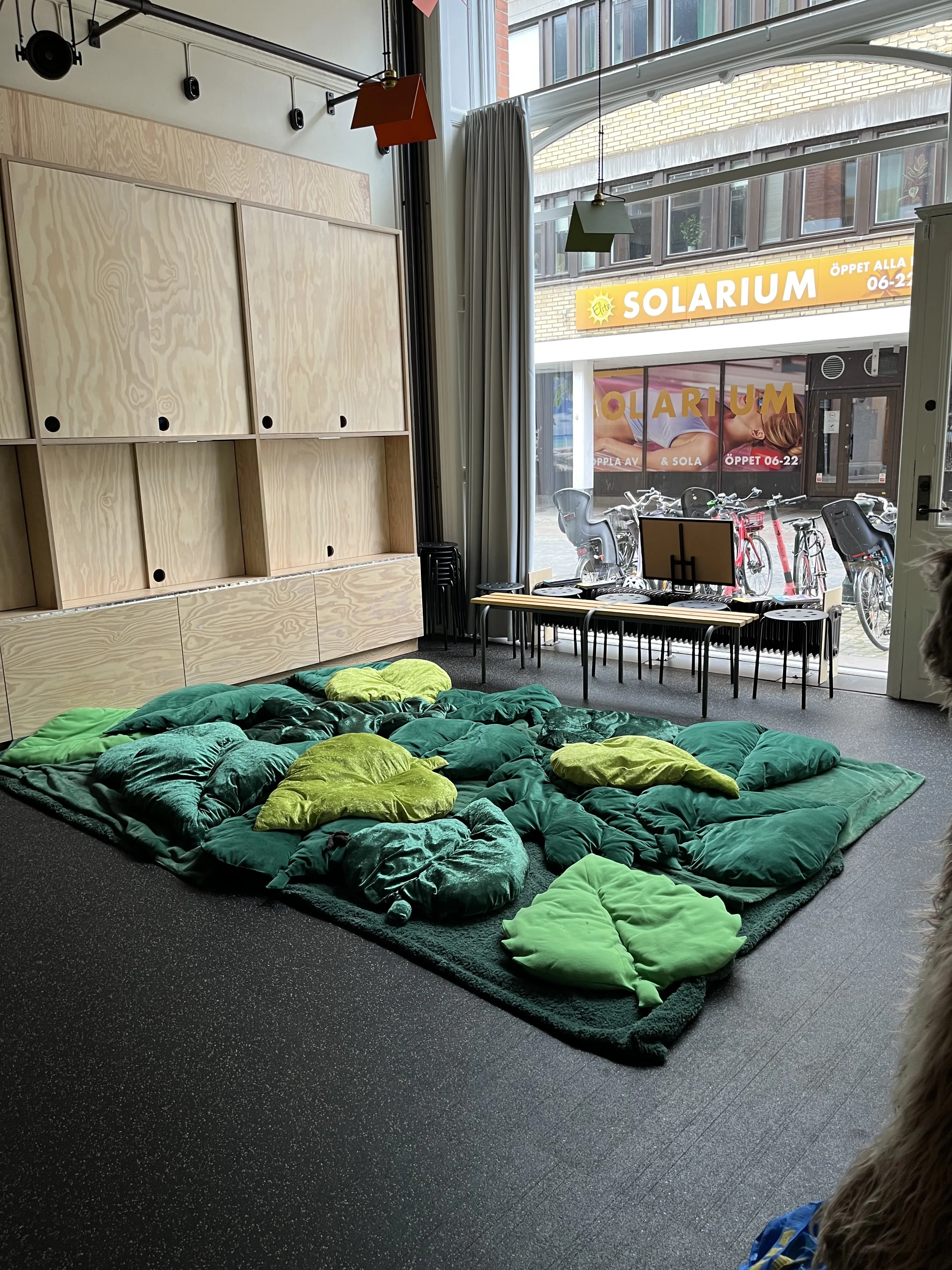 Gröna lövformade kuddar på golvet, en träbänk och några pallar. Ett stort fönster ut mot gatan.