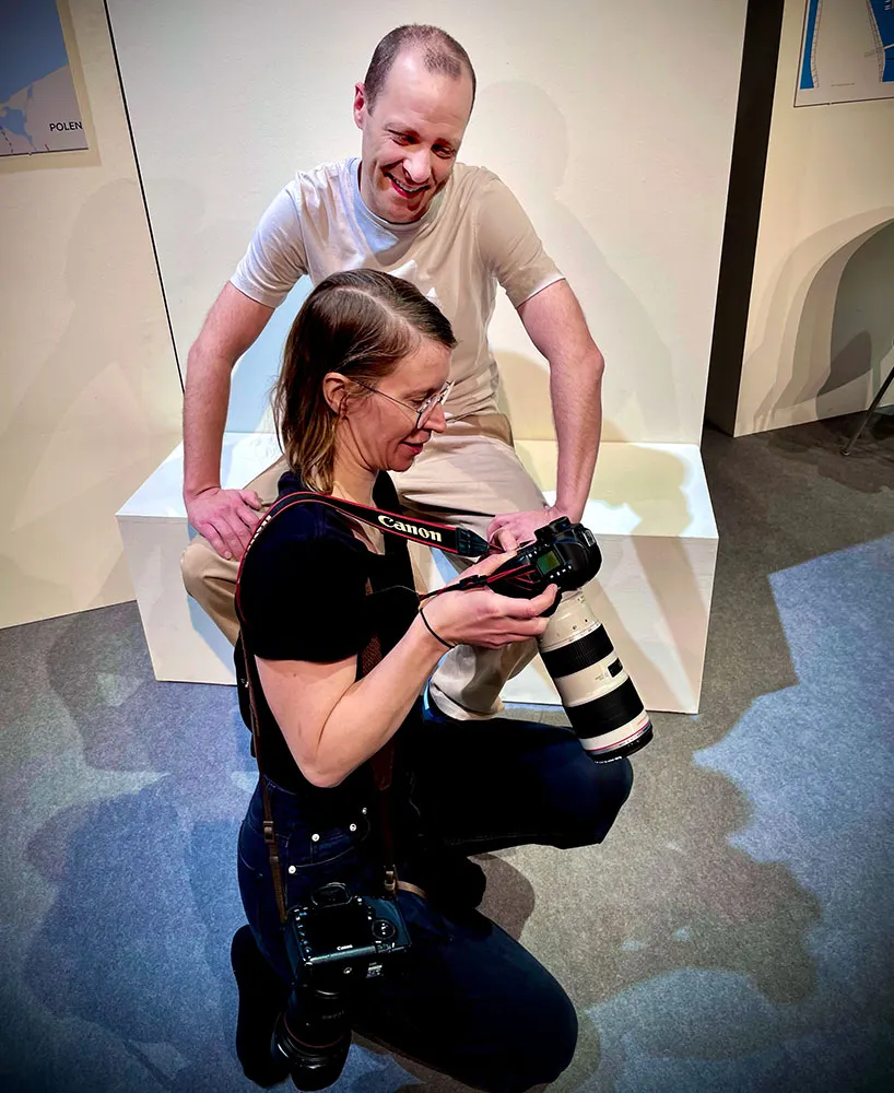 Föreställningsfotografen Emmalisa Pauly sitter på huk på scenen och visar skådespelaren Erik Borgeke bilder på kamerans display under fotografering av Underdog på Hippodromen 2023. Erik Borgeke sitter avslappnat på en vit scenografi-del och ler.