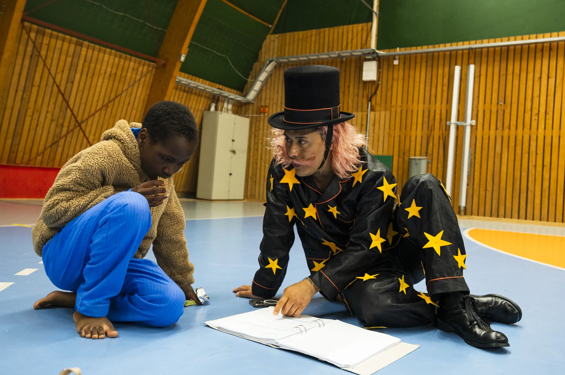 En skådespelare i stjärnmönstrad kostym och hög hatt sitter på golvet med ett barn. De tittar tillsammans i ett manus i en idrottshall.