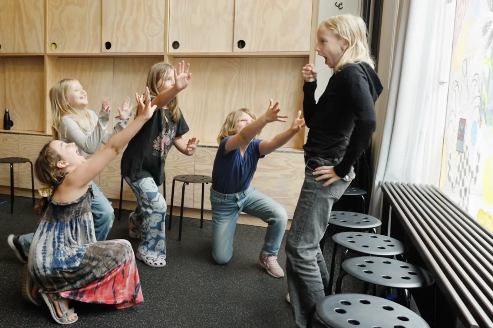 Teaterlek, några barn spelar upp en scen. Ett barn står upp och håller handen som en mikrofon. Runt om står andra barn med uppsträckta händer som vid en konsert.