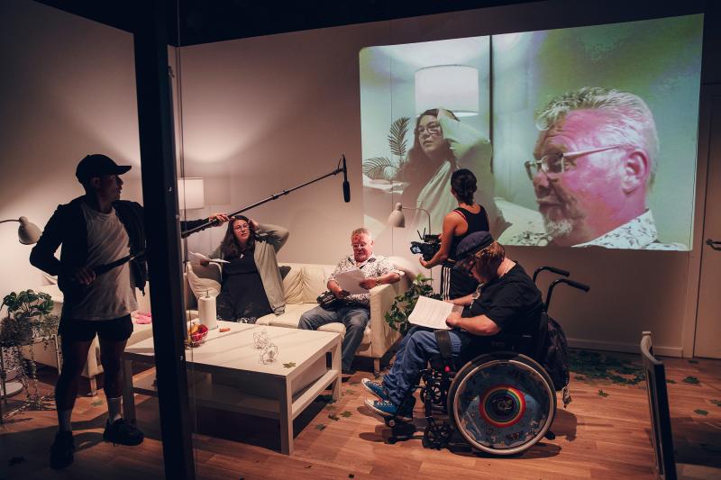 Foto: Emmalisa Pauly En grupp civila människor sitter och samtalar vid en soffgrupp och bredvid står skådespelare med mikrofoner som riktas mot de som talar.
