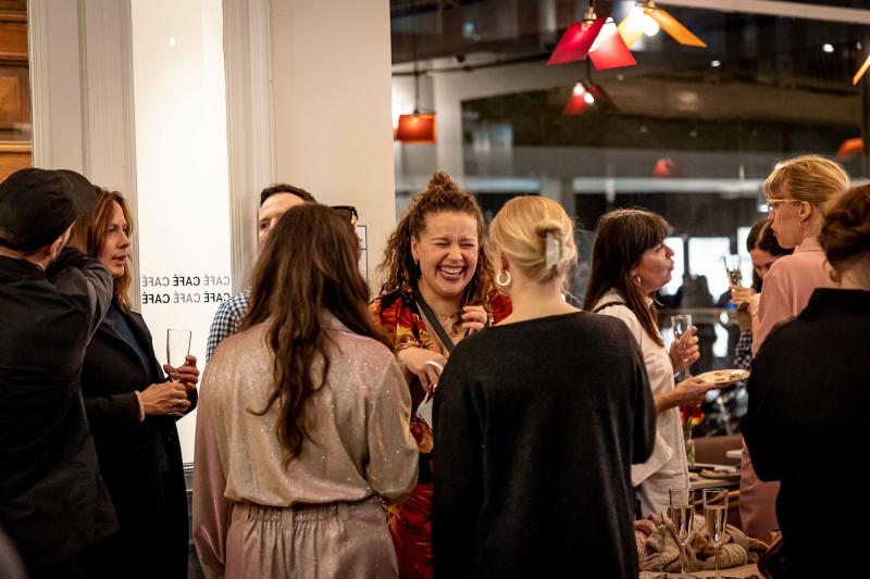 Jenny Baumgartner Folk som minglar i teatercaféet. En person i röd tröja står och skrattar tillsammans med två personer som vänder ryggen till mot kameran.