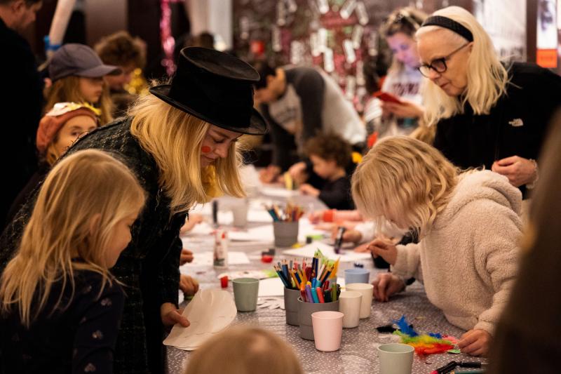 Jenny Baumgartner Pyssel vid långbord. En vuxen med hög svart hatt och clownröda kinder står tillsammans med barn som böjer sig över ett bord med papper, pennor, saxar och paljetter