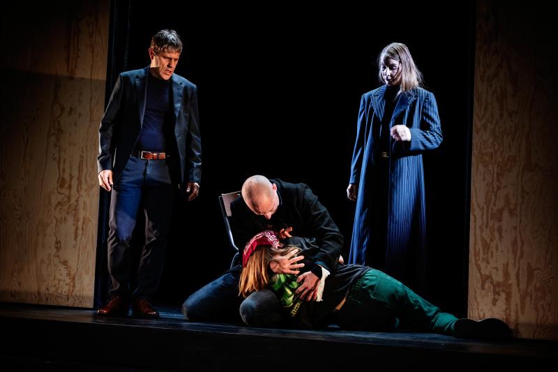 Foto: Emmalisa Pauly En dramatisk scen där en man i rock håller en person i sin famn som ligger på golvet med en röd sjal runt huvudet. Två andra personer, en man i kostym och en kvinna i randig kappa, står bredvid och ser på med bekymrade ansiktsuttryck.