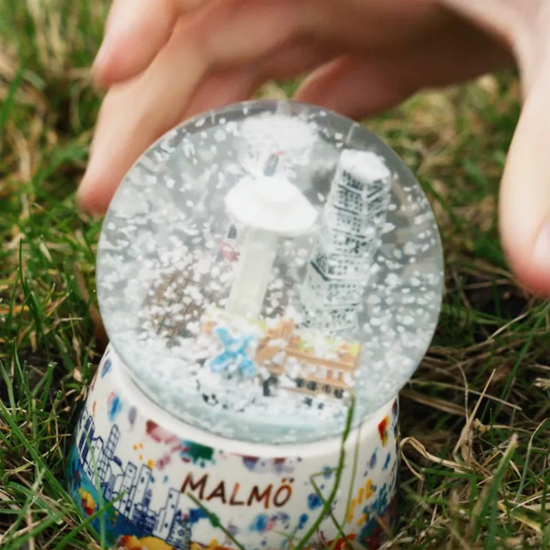 En skakad snöglob med Malmö som motiv står på en gräsmatta. Vi ser en hand som är på väg att greppa globen.
