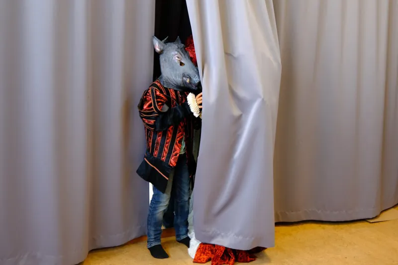 Ett barn med svartrödrandig kostymkappa och en mask i form av ett vildsvinshuvud står halvt gömd bakom en ridå