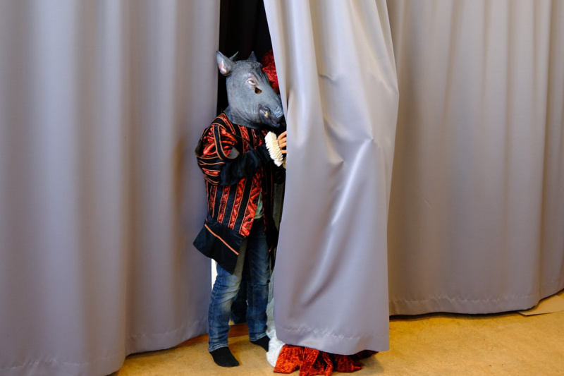 Ett barn med svartrödrandig kostymkappa och en mask i form av ett vildsvinshuvud står halvt gömd bakom en ridå