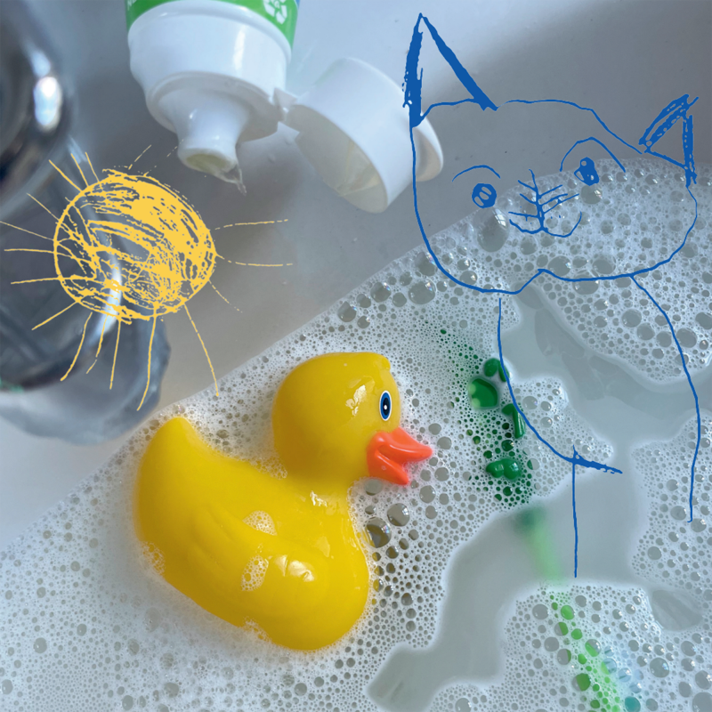 En gul plastanka ligger i ett skumbad tillsammans med vad som liknar en grön barntandborste. Ovanpå är ritade teckningar i form av en sol och en katt.I kant av bild syns tandkrämstub och kran.